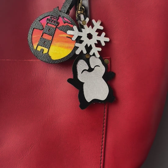 Handcrafted leather charm featuring a laughing penguin with a silver snowflake on top. The charm is designed in a sleek black and silver color combination and includes a lobster clasp for easy attachment to purses or use as a keychain.