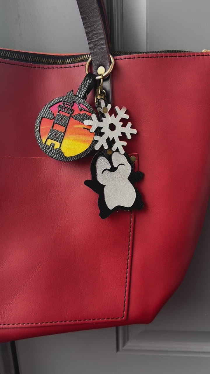 Handcrafted leather charm featuring a laughing penguin with a silver snowflake on top. The charm is designed in a sleek black and silver color combination and includes a lobster clasp for easy attachment to purses or use as a keychain.