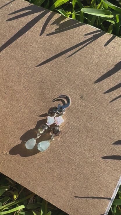 A pair of silver-colored three petal cathedral drop earrings with a gemstone drop, placed on a brown surface with a blurred background.