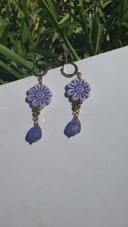 A pair of blue glass drop earrings with gold plated brass findings, displayed on a flat surface with grass and a clear sky in the background.