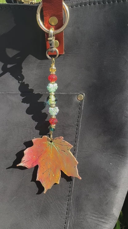 A handcrafted maple leaf charm made of vegetable tan leather with a brass hardware and multicolored beads, designed to be worn on a bag.