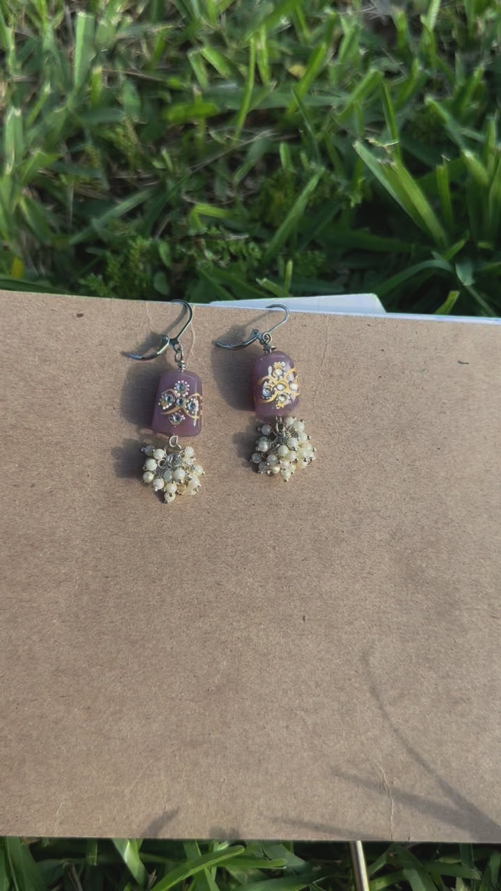 Silver earrings with a mauve color cluster of pearls and a hand painted glass bead, displayed on a spiral notebook page with grass and shadows in the background.