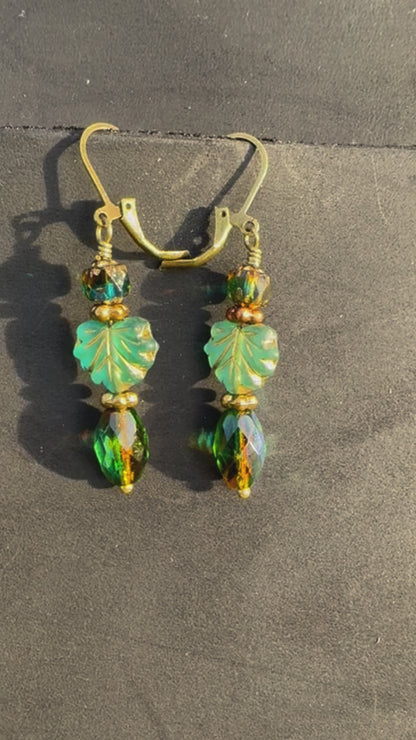 A pair of handcrafted earrings with green and blue czech glass beads, antique bronze finish, and brass hardware on a textured background.