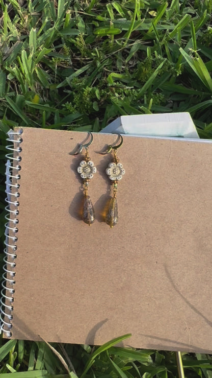A pair of white hibiscus gold drop earrings with gold plated brass findings, displayed on a notebook.