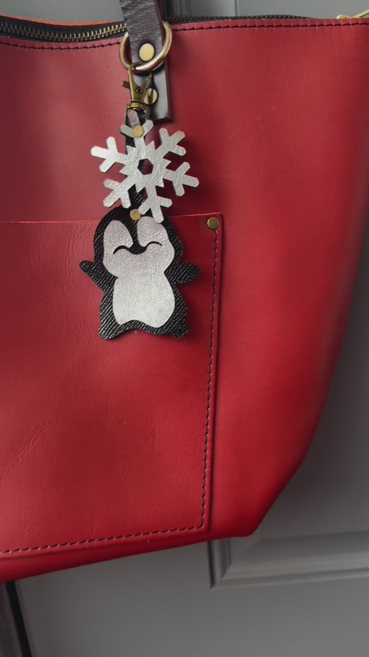 Handcrafted leather charm featuring a laughing penguin with a silver snowflake on top. The charm is designed in a sleek black and silver color combination and includes a lobster clasp for easy attachment to purses or use as a keychain.