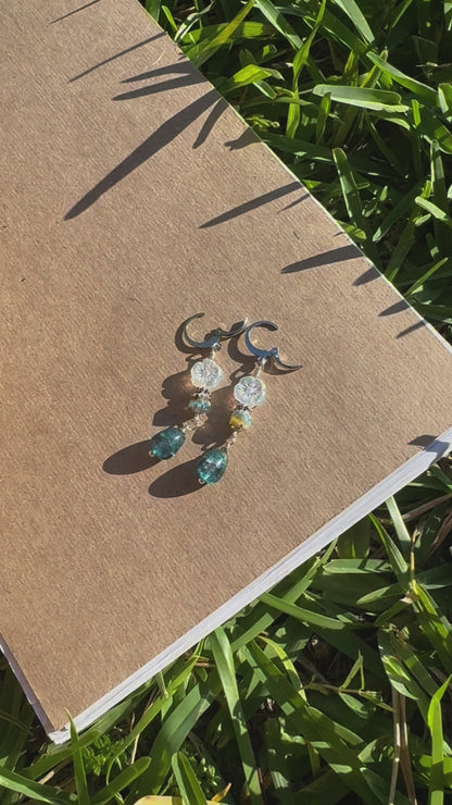 A pair of silver drop earrings with blue and clear embellishments, displayed on a brown notebook outdoors.