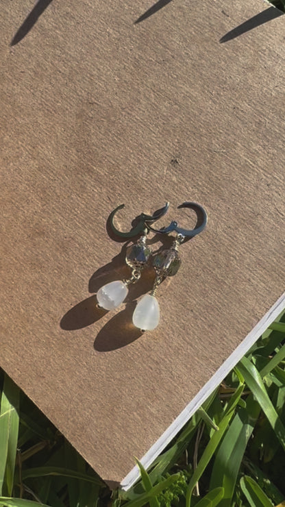 A pair of pale blue silver drop earrings with a cathedral-like design, displayed on a brown paper surface, with blurred greenery in the background.
