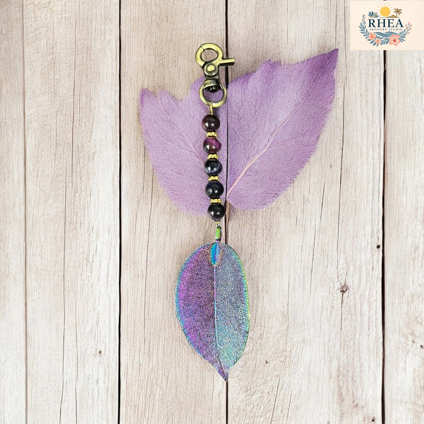 Decorative keychain with purple and green leaves on a wooden surface, featuring the brand 'RHEA'.