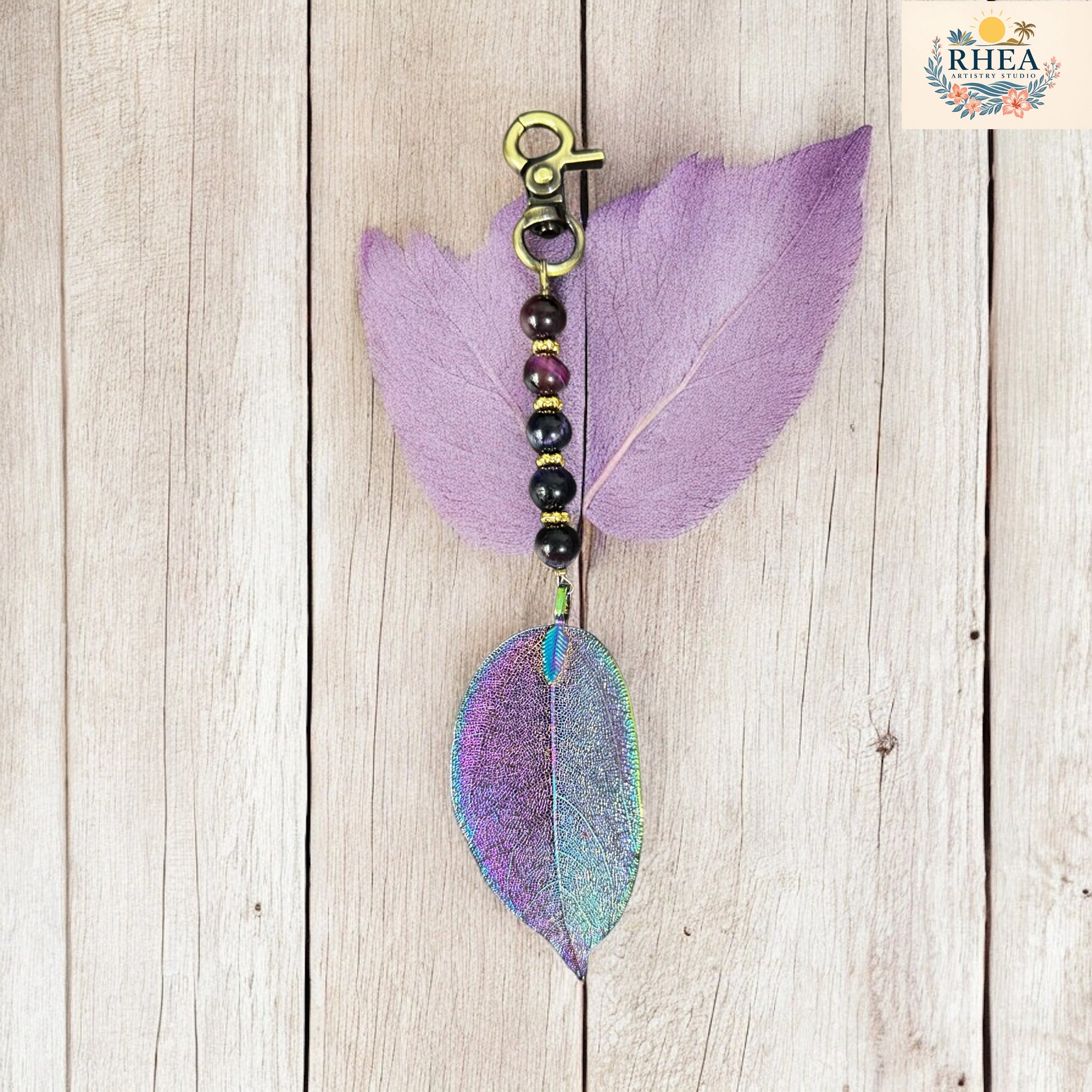 Decorative keychain with purple and green leaves on a wooden surface, featuring the brand 'RHEA'.