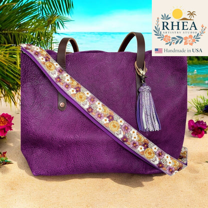 Purple tote bag with floral purse strap and tassel on a beach, featuring the RHEA Artistry Studio logo.