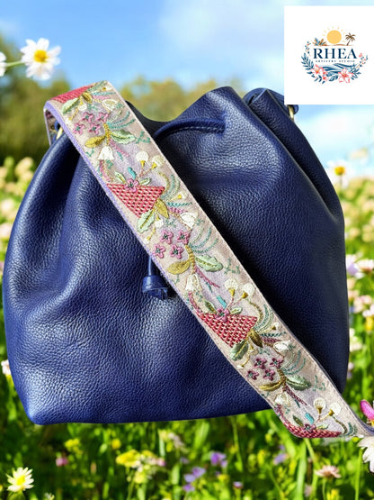 Embroidered crossbody strap with lavender background featuring pink basket motifs, pastel green vines, and small purple and yellow florals, shown on a navy leather bag.
