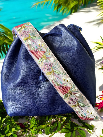 Embroidered crossbody strap with lavender background featuring pink basket motifs, pastel green vines, and small purple and yellow florals, shown on a navy leather bag on a sandy beach