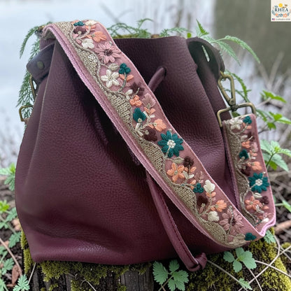 A burgundy pebbled leather bucket bag with a wide embroidered shoulder strap, resting on a mossy log outdoors.