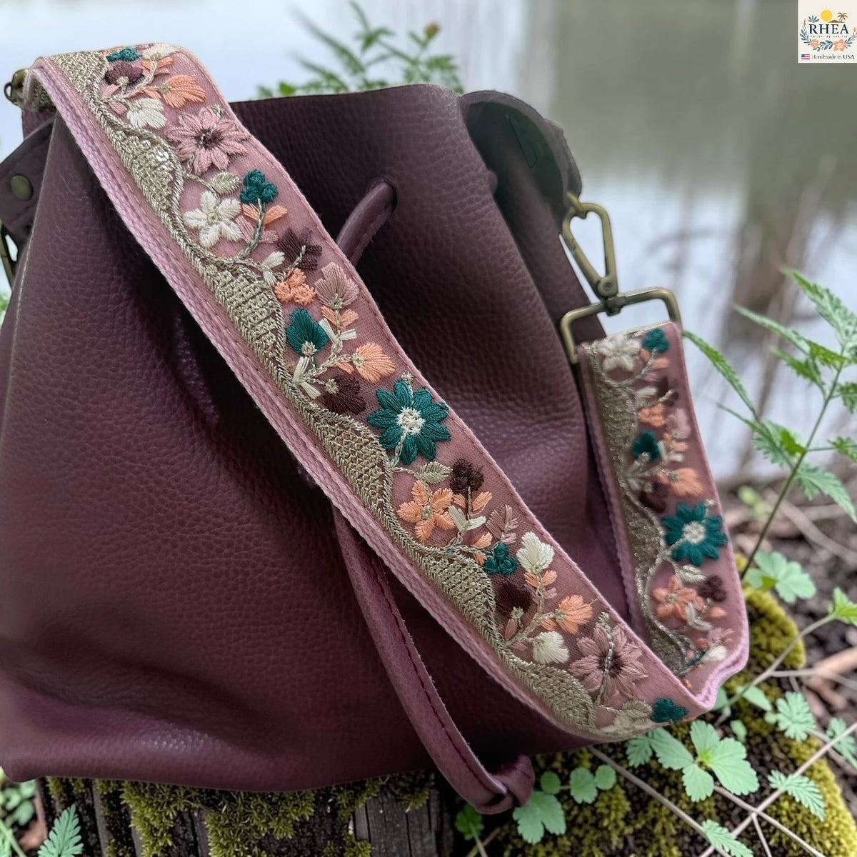 A dark plum leather bucket bag with a wide, adjustable embroidered shoulder strap, resting outdoors on a mossy log with green foliage in the background.