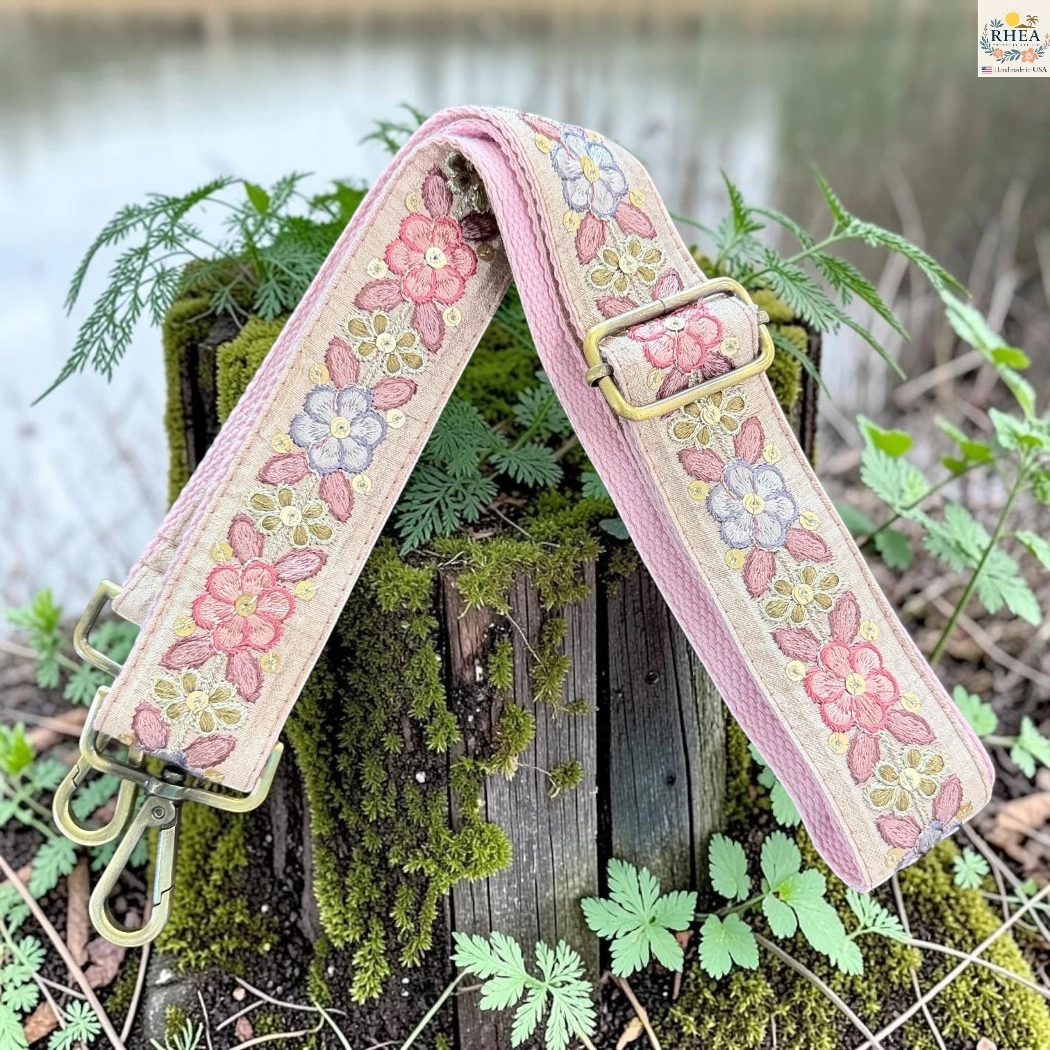 A floral embroidered strap with a pink border, resting on a mossy wooden stump outdoors near water. The strap features pink, purple, and yellow flowers on a beige background with brass-tone metal hardware.