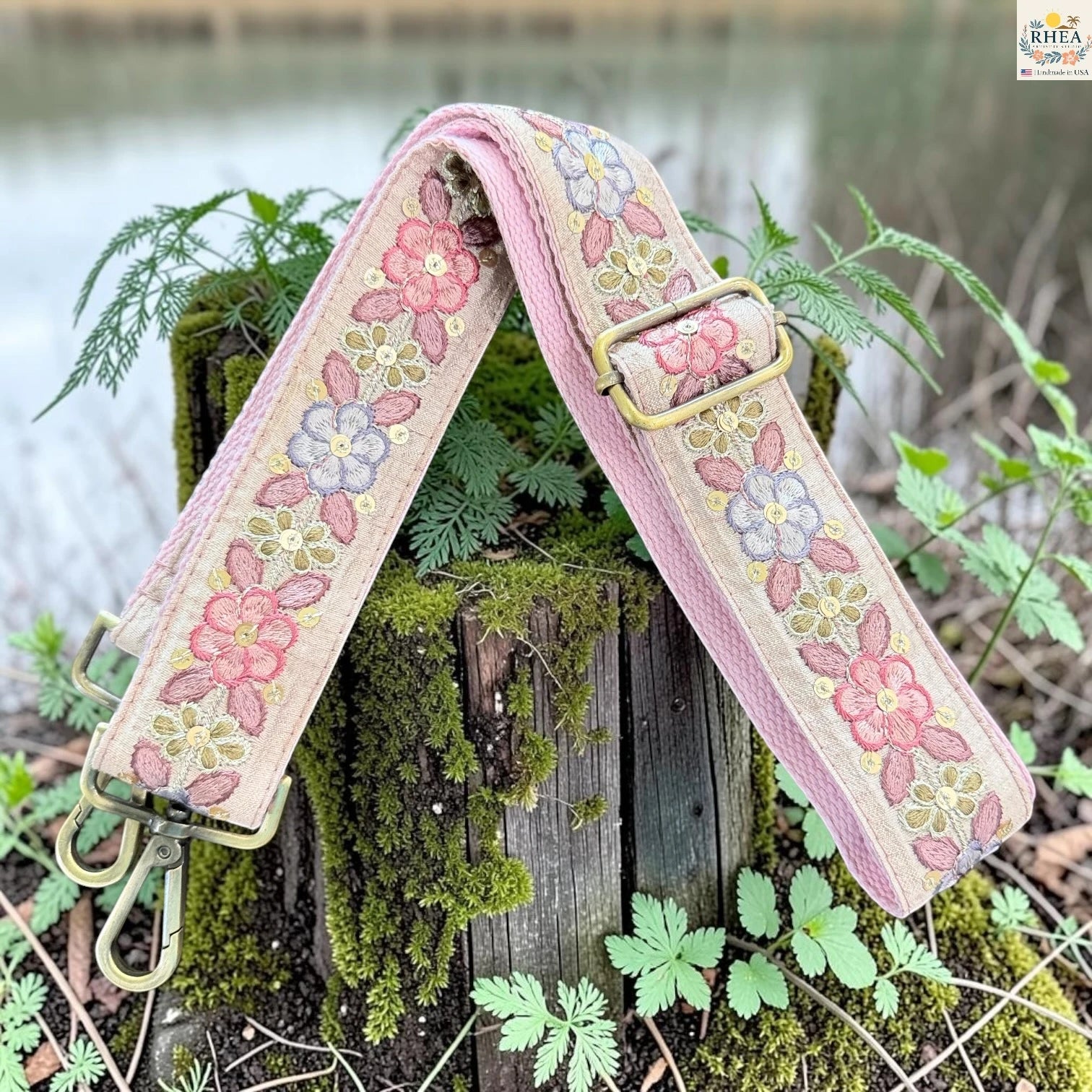A floral canvas shoulder strap with pink, blue, and yellow embroidered flowers, resting on a mossy wooden stump outdoors.