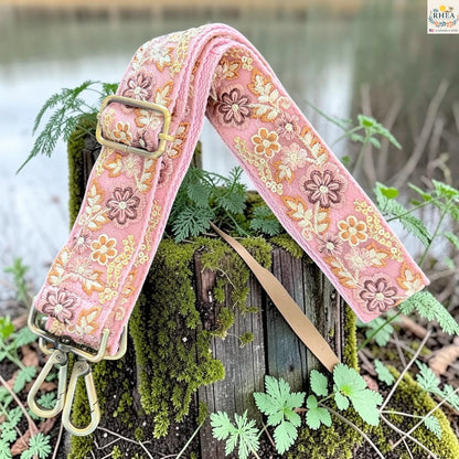 A pink floral embroidered fabric camera strap with brass hardware, draped over a mossy wooden post outdoors near water.