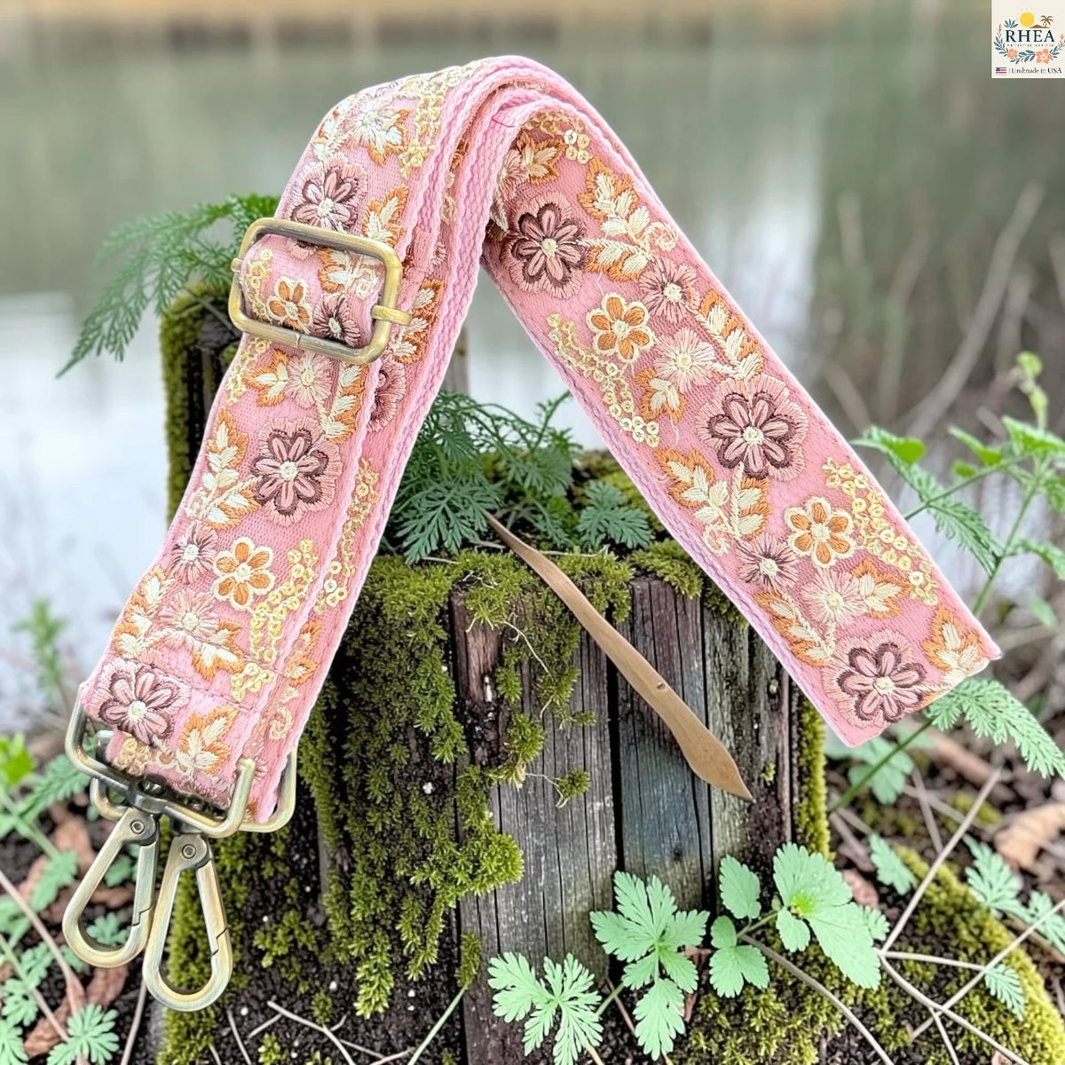 A pink floral fabric camera strap with brass hardware, draped over a mossy wooden stump outdoors near green plants and water.