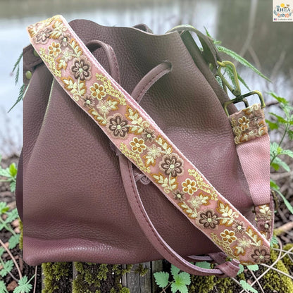 A dusty rose pebbled leather shoulder bag with a wide, decorative floral embroidered strap, resting outdoors on a mossy log with green foliage and water in the background.
