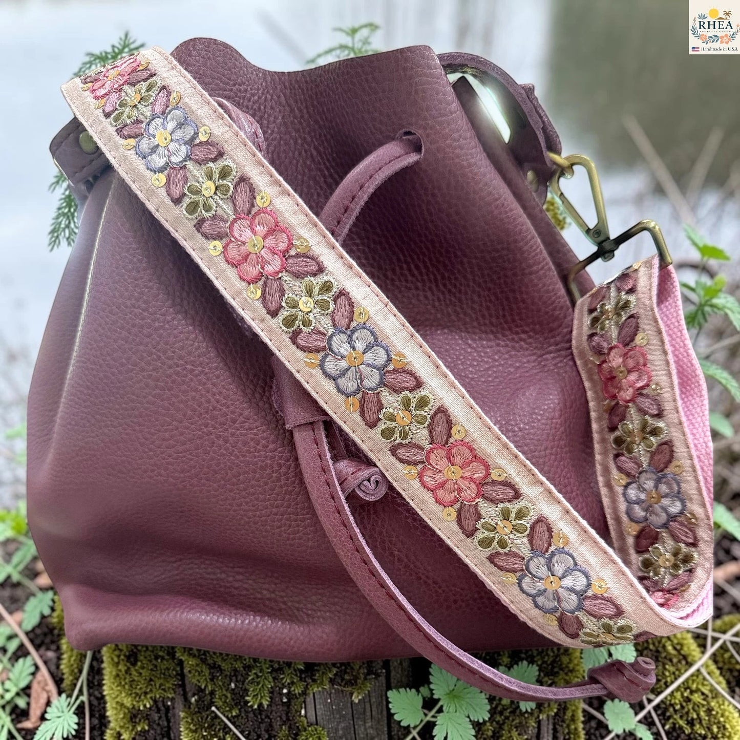 A dusty rose pebbled leather bucket bag with a wide, adjustable embroidered shoulder strap, resting on a mossy log outdoors.
