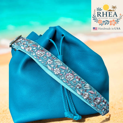 Blue drawstring bag with floral purse strap on a beach, featuring RHEA Artistry Studio logo.