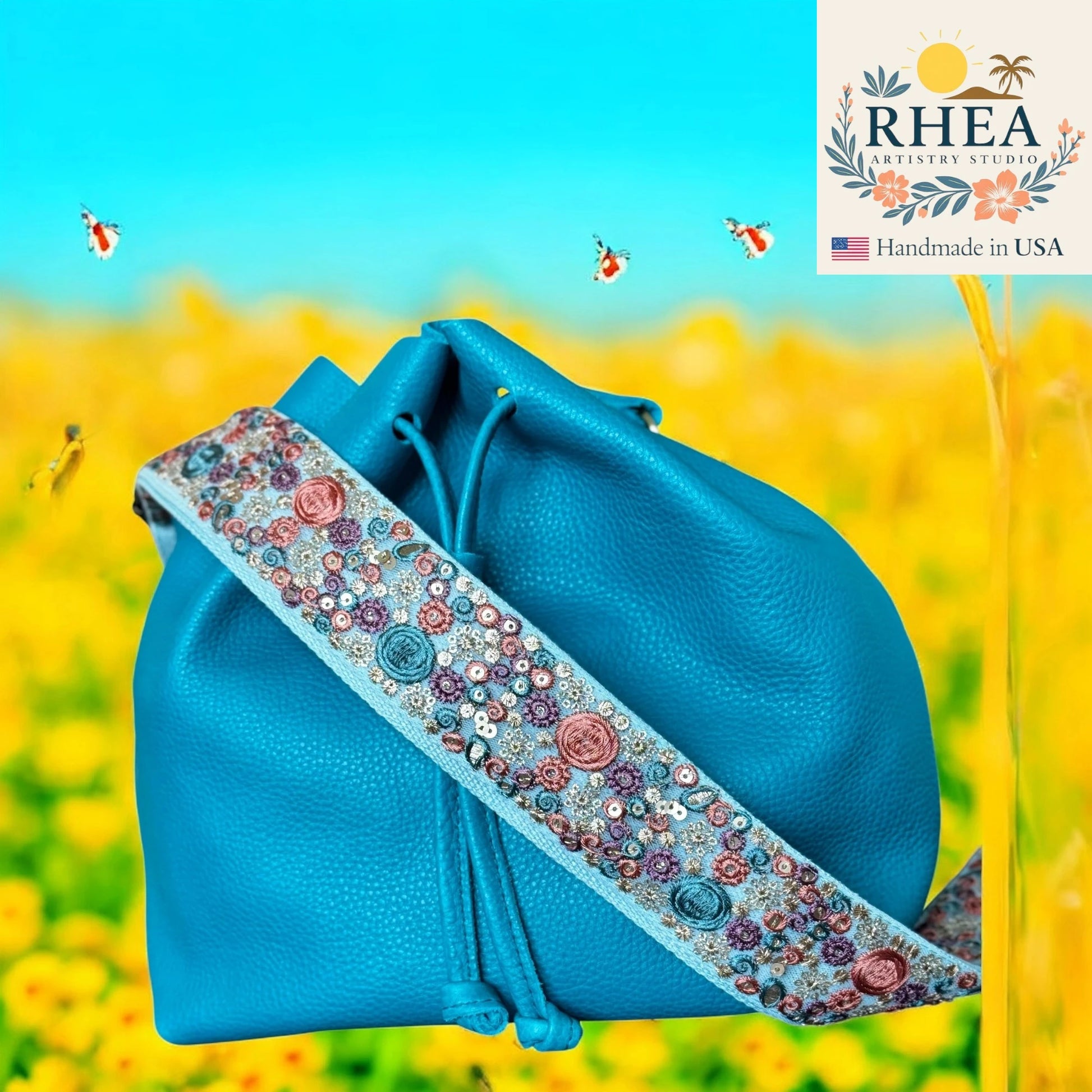 Blue bag with decorative purse strap against a yellow flower field background, featuring the RHEEA brand.