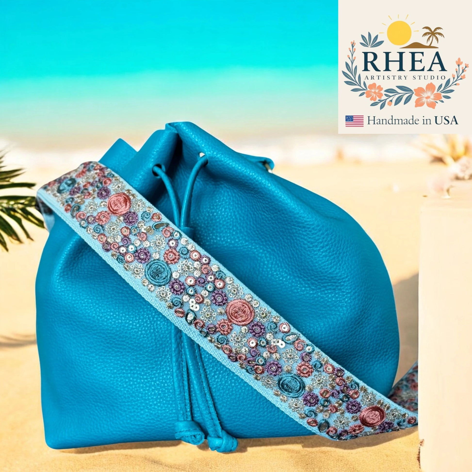 Blue bag with a floral purse strap on a beach background, featuring the RHEA brand.