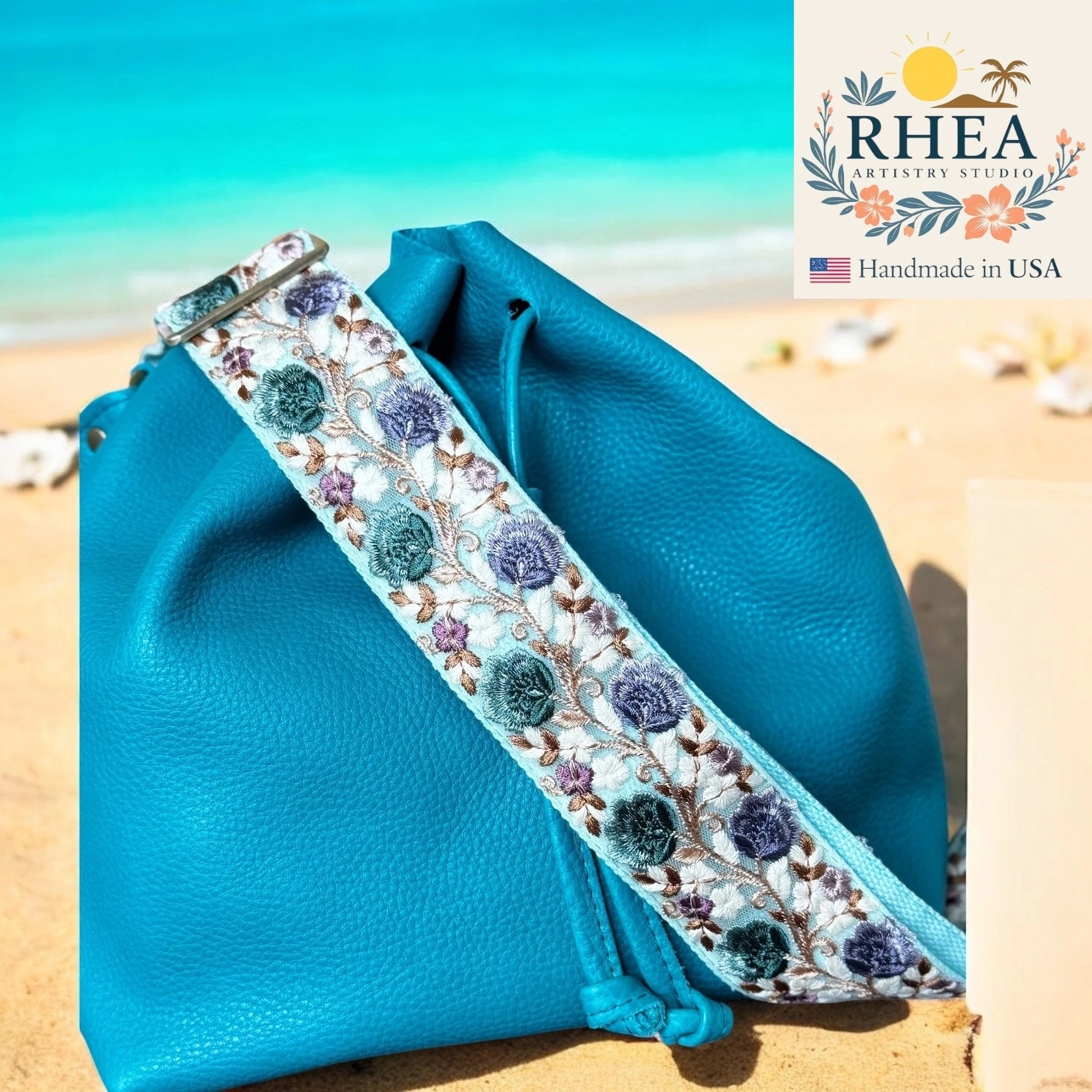 Blue pouch with floral purse strap on a beach, featuring RHEA Artistry Studio logo.