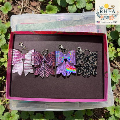 Rhea Artistry Studio handmade animal print bow bag charms in pink glitter, zebra, purple holographic, and leopard patterns with brass hardware.