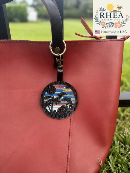 Rhea Artistry Studio hand-painted leather bag charm featuring dolphins, palm trees, and a beach sunset scene. Black textured leather accent.
