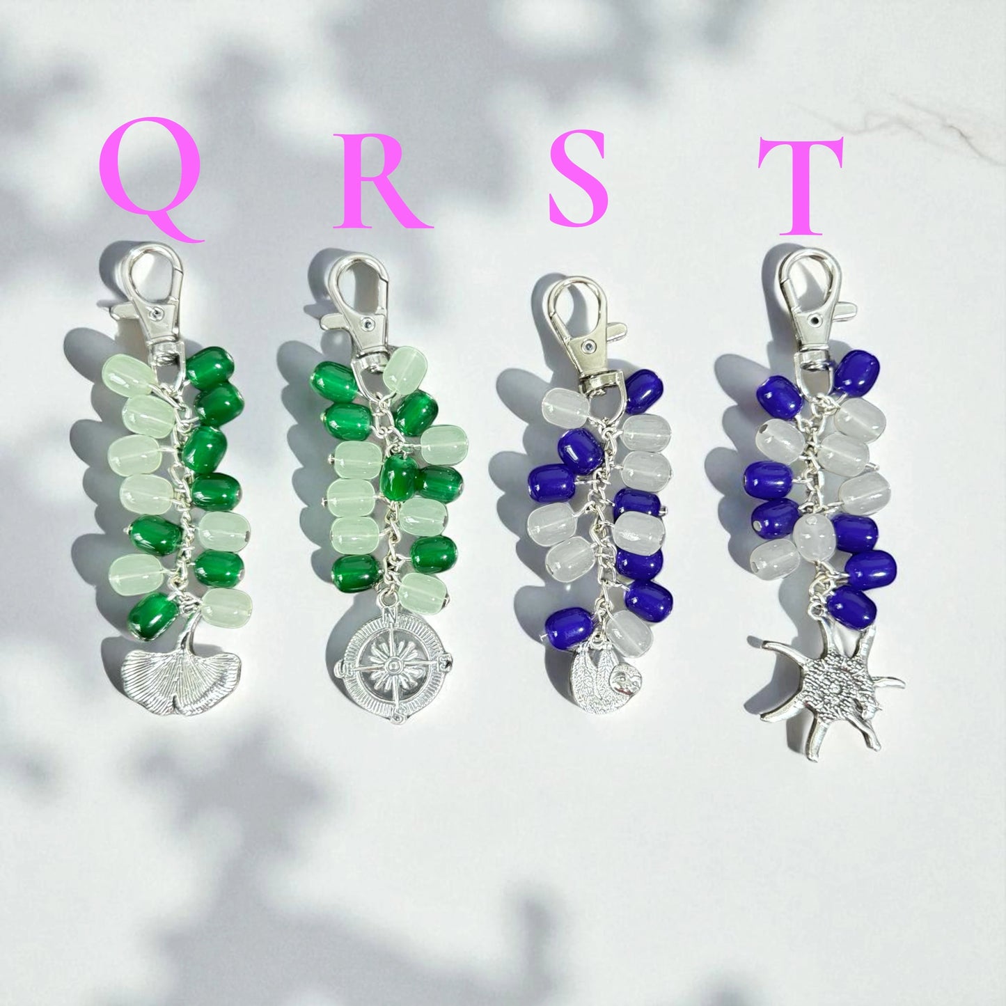 Rhea Artistry Studio handmade beaded keychains in green, blue, and white. Features silver-tone charms: ginkgo leaf, compass, sloth, and sunburst.