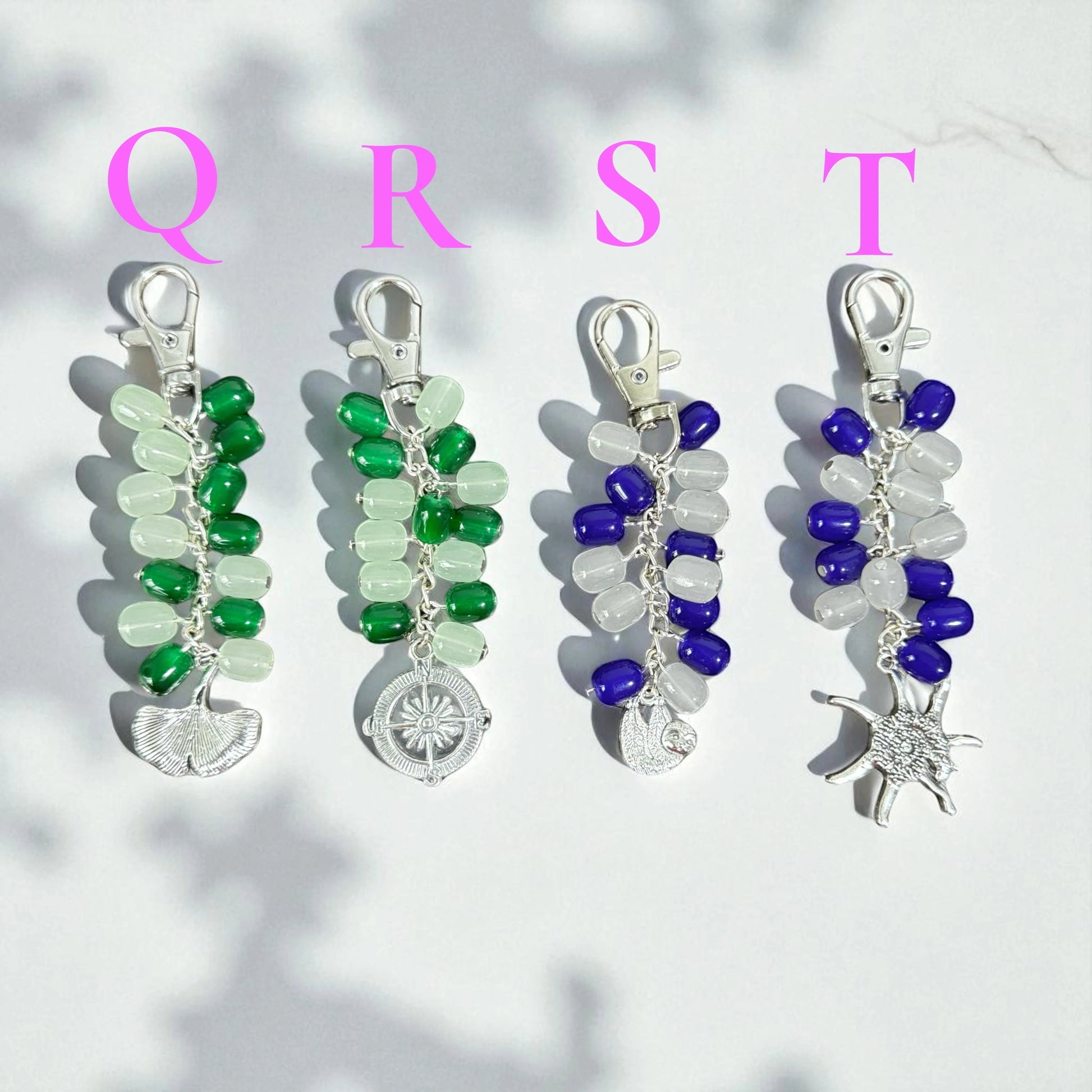 Rhea Artistry Studio handmade beaded keychains in green, blue, and white. Features silver-tone charms: ginkgo leaf, compass, sloth, and sunburst.