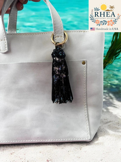 Rhea Artistry Studio black leather tassel bag charm with iridescent shimmer and gold lobster clasp.