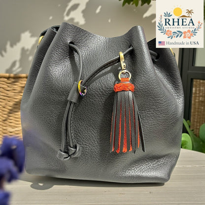 Rhea Artistry Studio handmade black leather tassel bag charm with orange accents and antique brass clasp.
