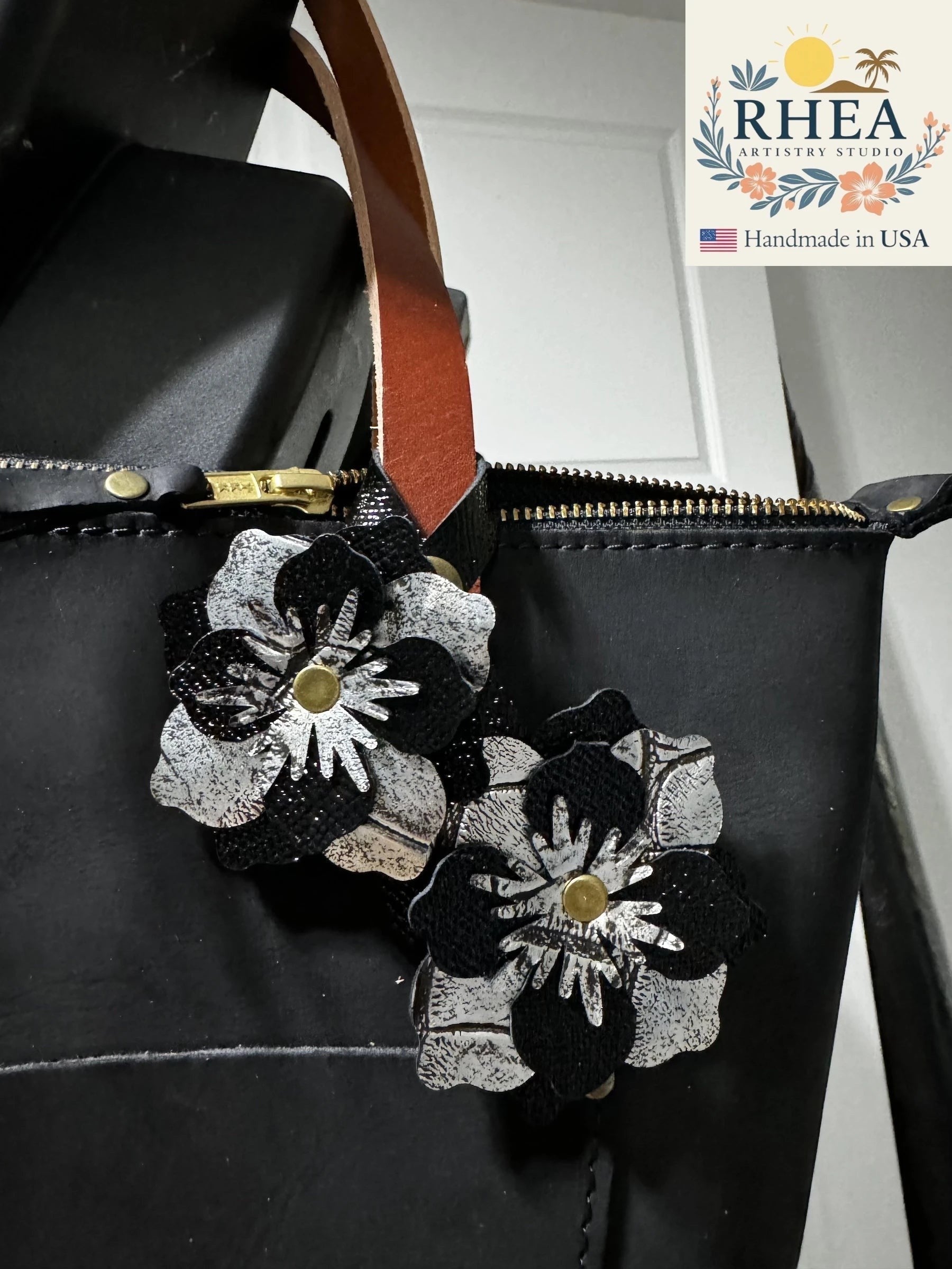 Rhea Artistry Studio black and silver leather flower double charm with brass rivets and snap fastener attached to a black leather bag.