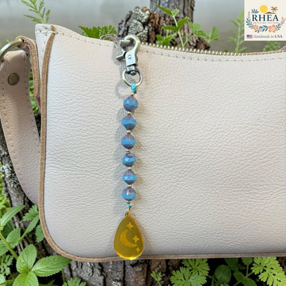 Rhea Artistry Studio blue Czech glass bead bag charm with brass accents and a yellow teardrop pendant featuring crescent moons and stars.