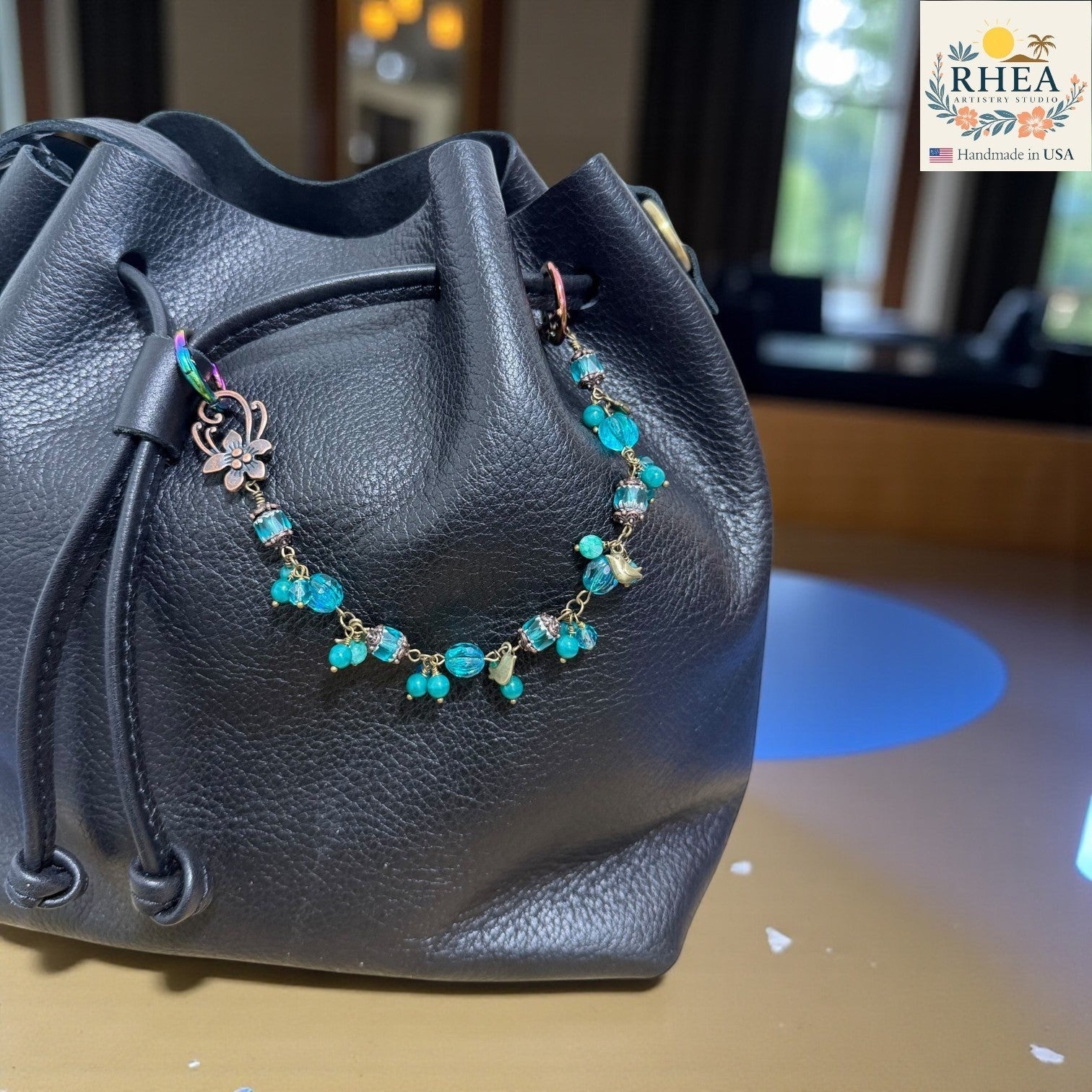 Rhea Artistry Studio handmade beaded purse charm with blue Czech glass beads and brass celestial charms attached to a black leather bag.