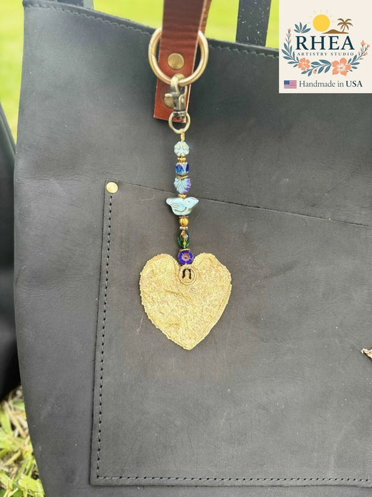 Rhea Artistry Studio blue bird charm with gold heart pendant and blue beads, attached to a black leather tote bag.