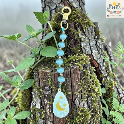 Rhea Artistry Studio blue Czech glass bead bag charm with brass accents and hand-painted moon and stars pendant.