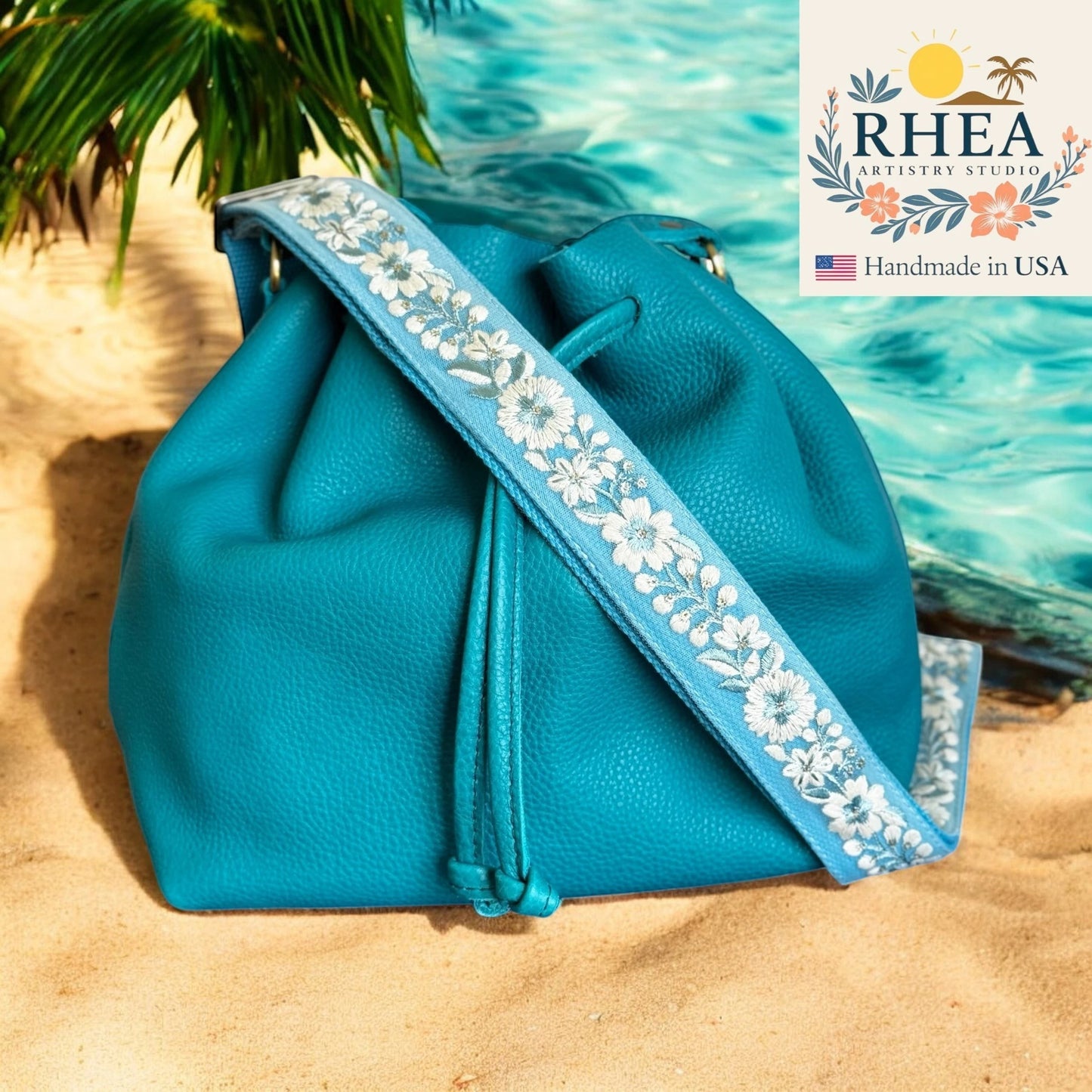 Rhea Artistry Studio blue crossbody strap with white floral embroidery, 1.5-inch wide, adjustable length, on a teal bucket bag.