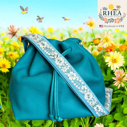Rhea Artistry Studio blue crossbody strap with white floral embroidery and brushed brass hardware, shown on a teal bucket bag.