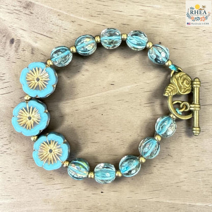 Rhea Artistry Studio handmade bracelet with blue floral glass beads and brass toggle clasp. Features clear, aqua, and gold beads.