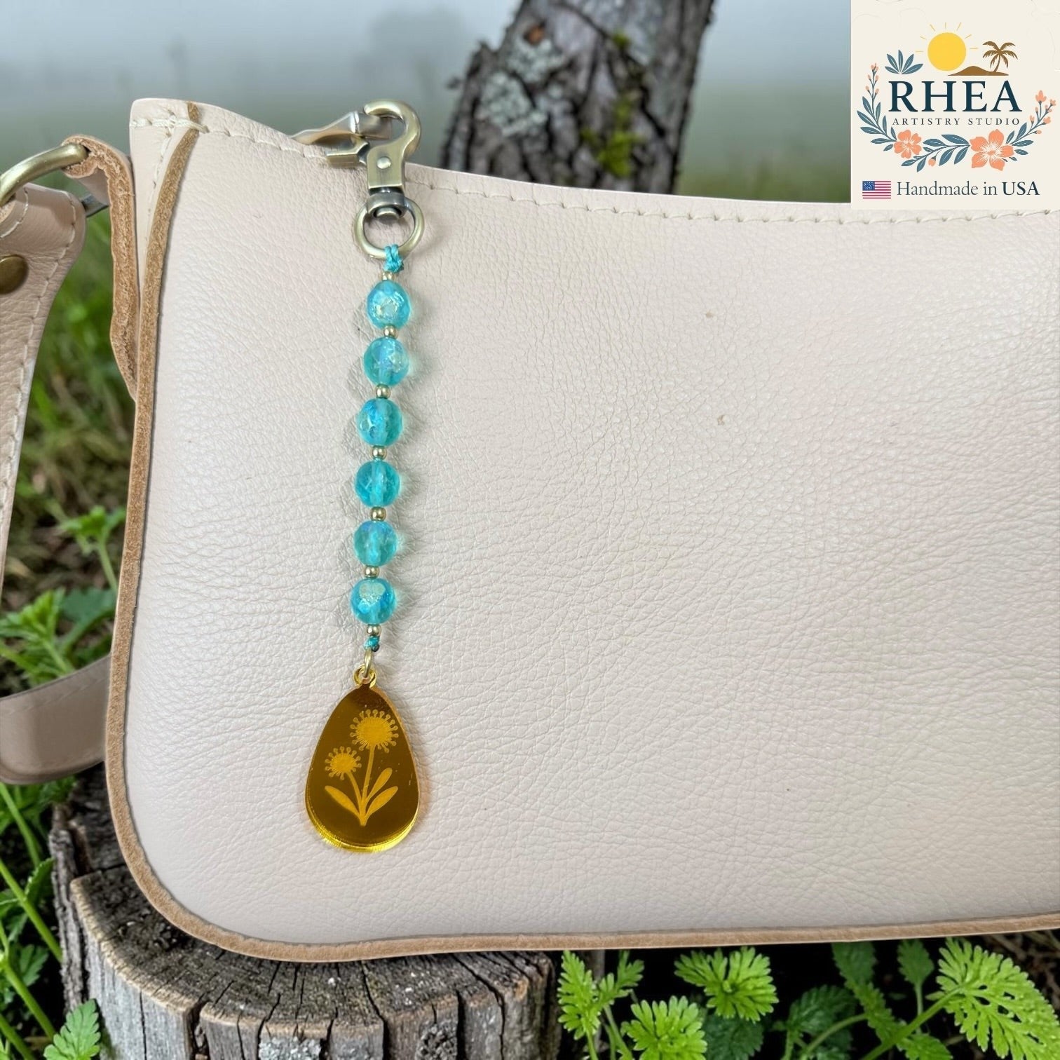 Rhea Artistry Studio bag charm with blue faceted glass beads and a gold engraved pendant featuring flowers.