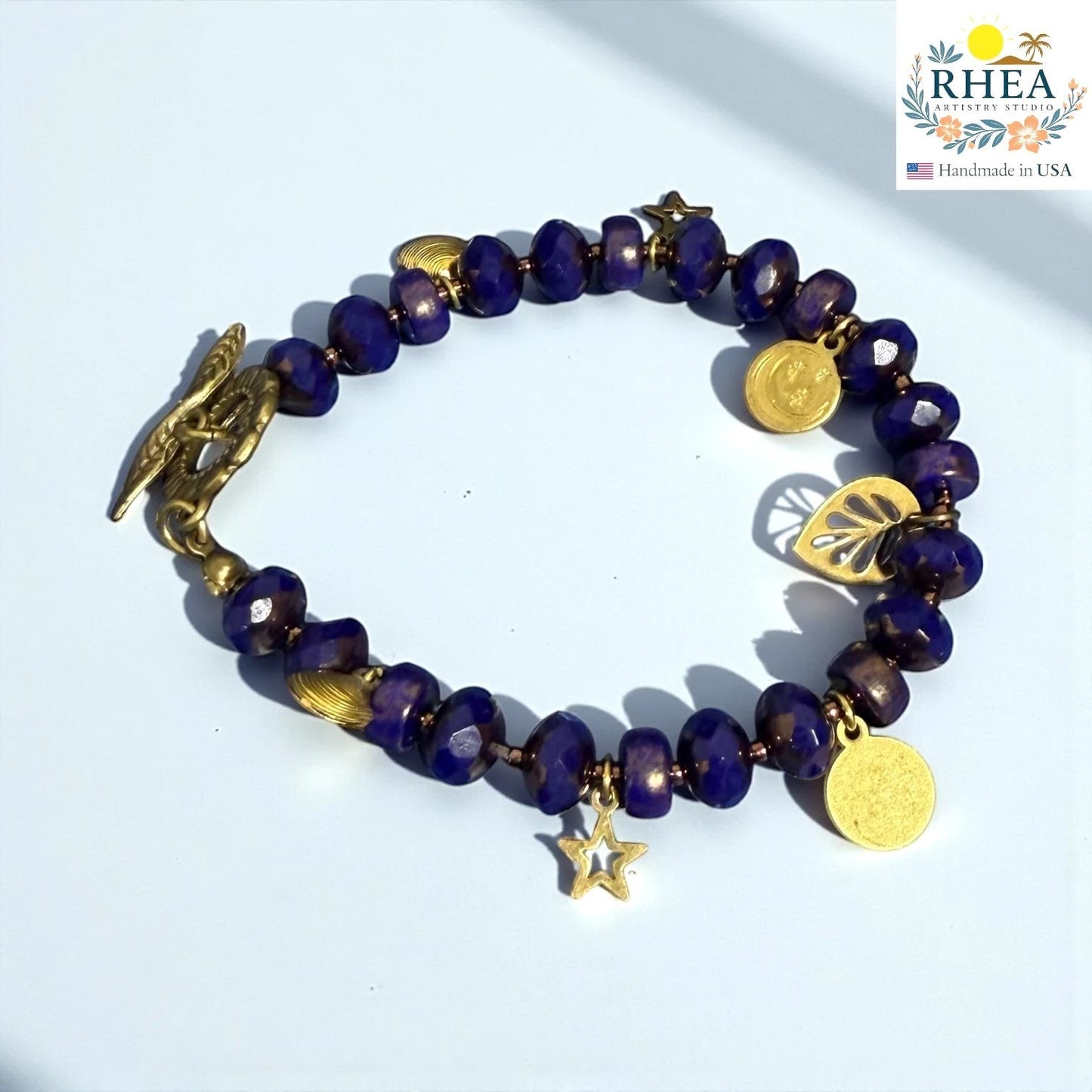 Rhea Artistry Studio handmade bracelet with deep blue faceted glass beads, gold star, moon, and leaf charms, and a floral toggle clasp.