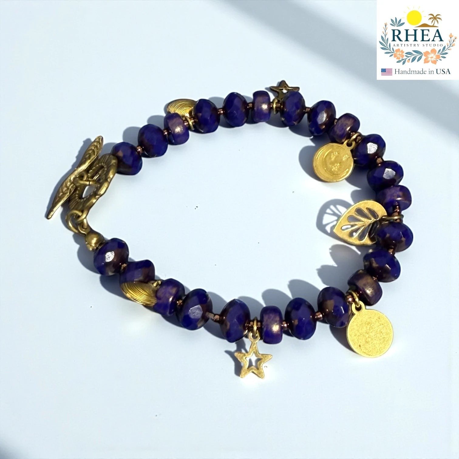 Rhea Artistry Studio handmade bracelet with deep blue faceted glass beads, gold star, moon, and leaf charms, and a floral toggle clasp.