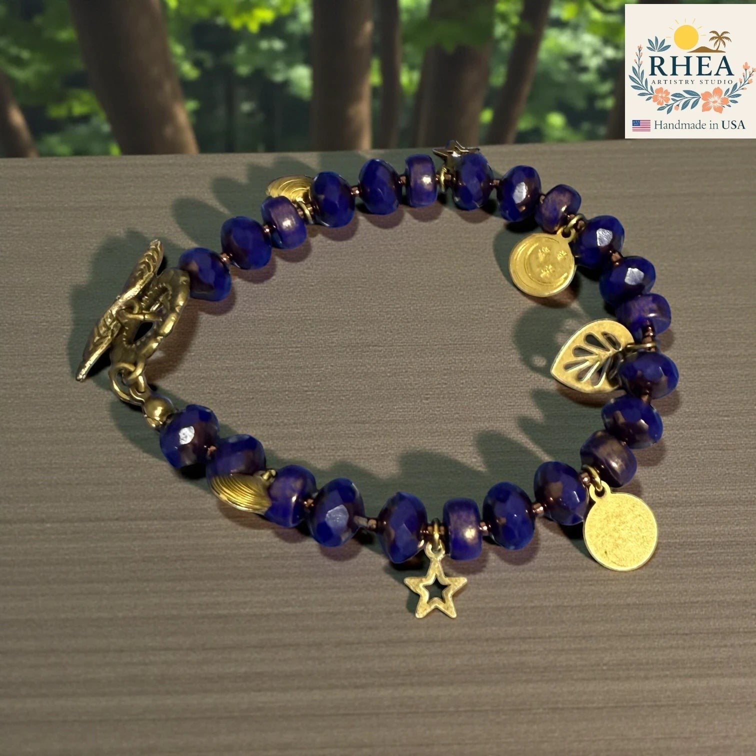 Rhea Artistry Studio handmade celestial bracelet with deep blue glass beads, brass star, moon, leaf charms, and floral toggle clasp.