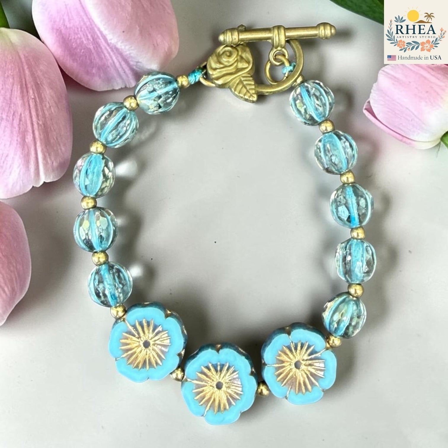 Rhea Artistry Studio handmade bracelet with blue glass beads, gold accents, and floral charms. Features a brass toggle clasp.