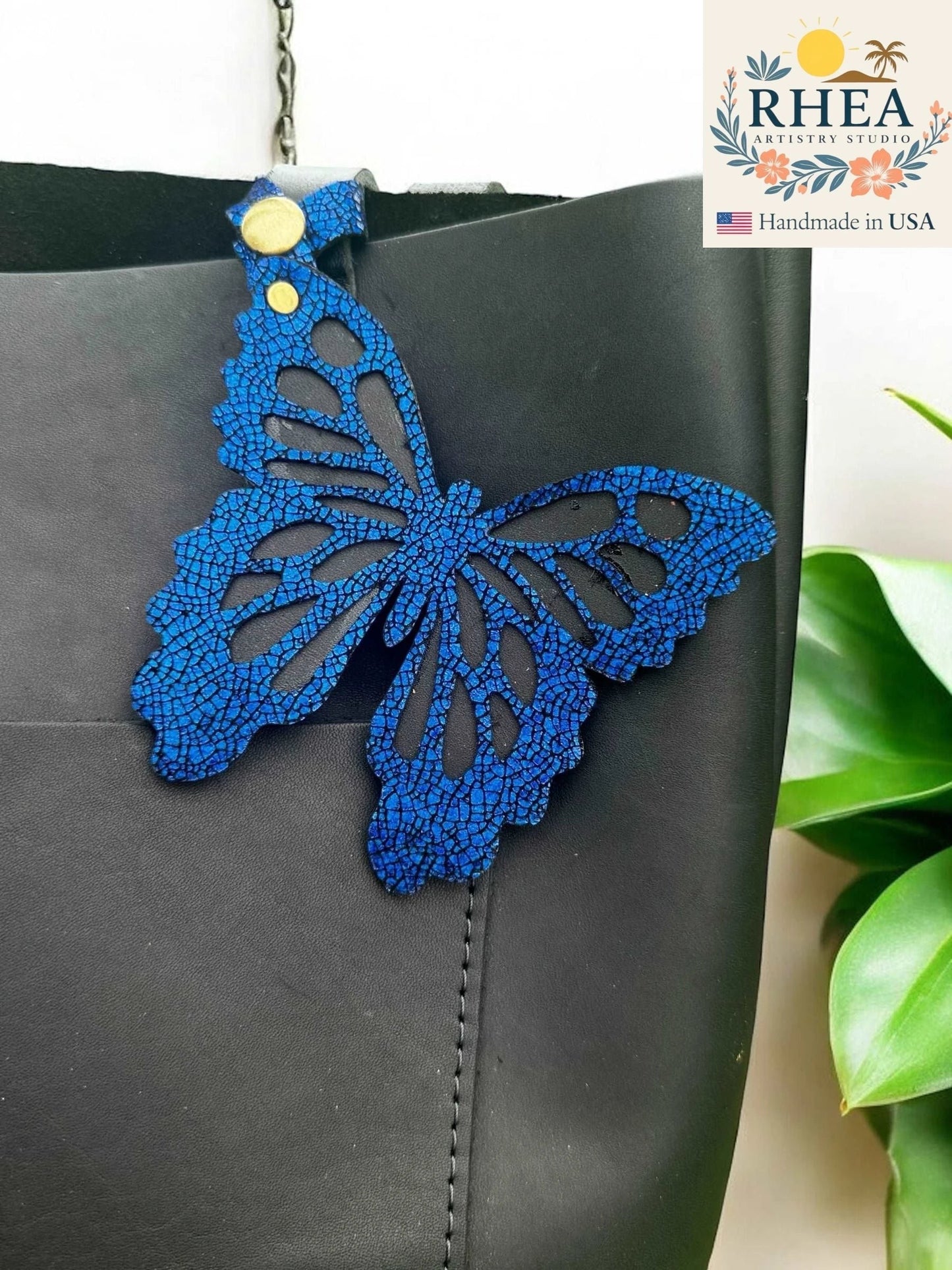 Rhea Artistry Studio blue leather butterfly charm with metallic iridescent finish, attached to a black bag.