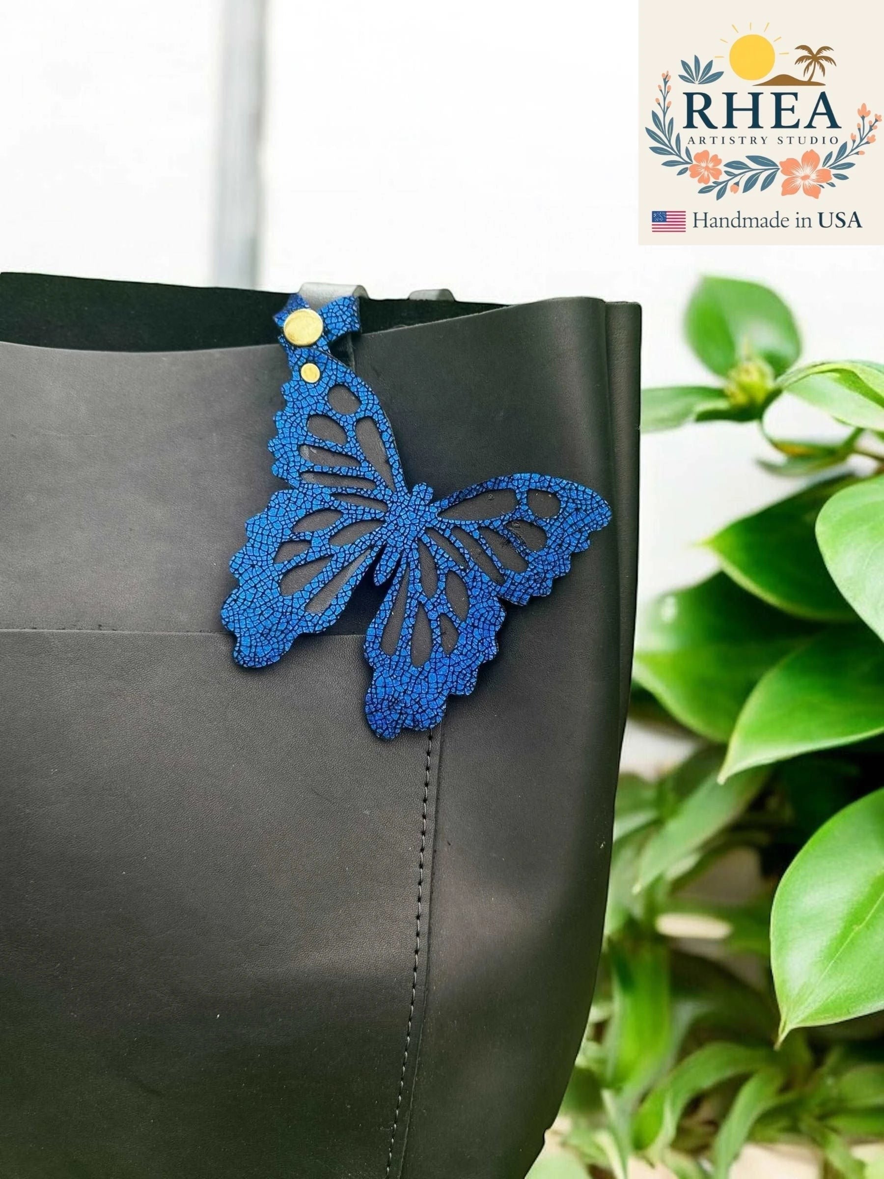 Rhea Artistry Studio blue leather butterfly charm with metallic iridescent finish and gold snap closure attached to a black tote bag.