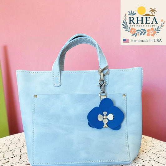 Rhea Artistry Studio blue leather orchid keychain charm with brushed brass hardware attached to a light blue tote bag.